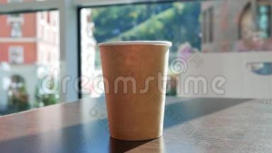 年轻女子从桌子上拿起咖啡馆里的纸杯茶。 喝茶，把杯子放在桌子上。 窗外的倒影